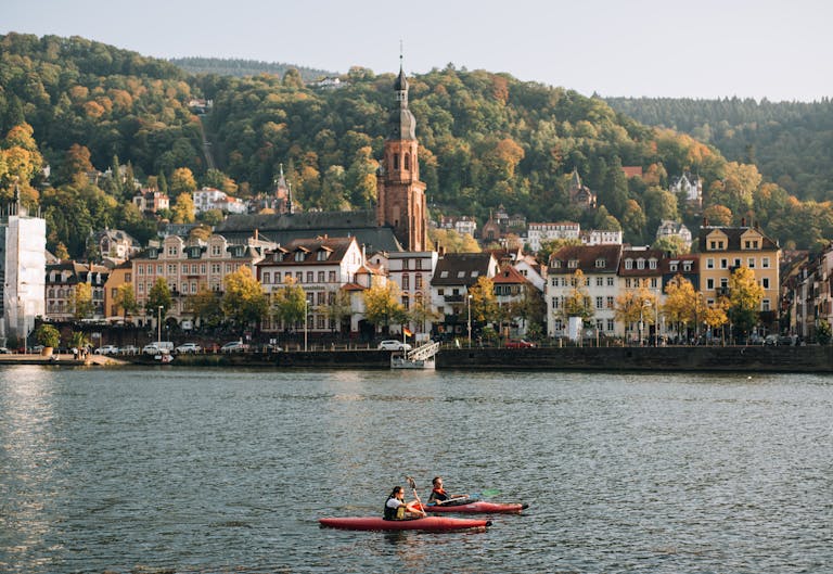 Kayakers on the Neckar River with Heidelberg's charming architecture and lush hills in the background.