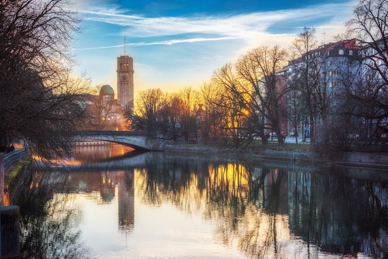 Scenic sunset view of Munich's riverside with a bridge, buildings, and trees reflecting in the water.