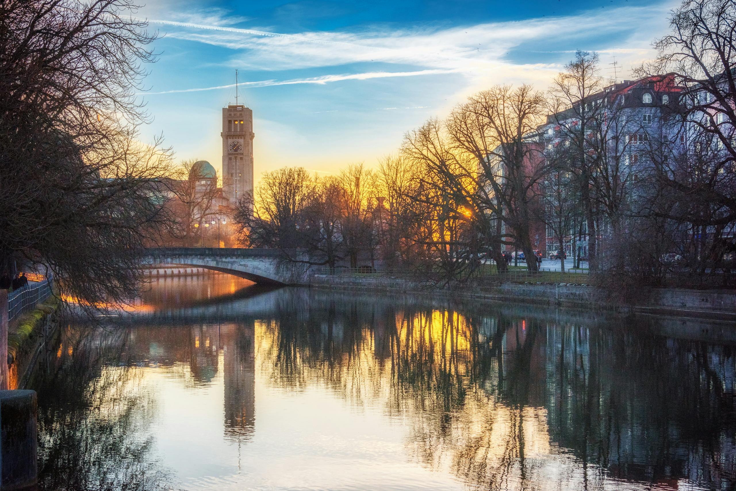 Scenic sunset view of Munich's riverside with a bridge, buildings, and trees reflecting in the water.